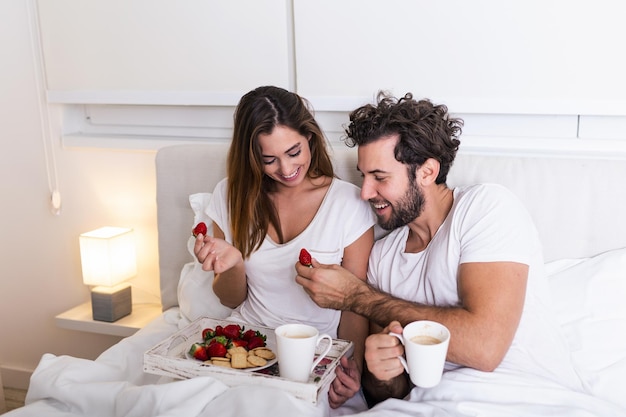 Casal bonito tomando café da manhã na cama no quarto. linda mulher alimentando o namorado com morangos na cama enquanto toma café da manhã e café no quarto