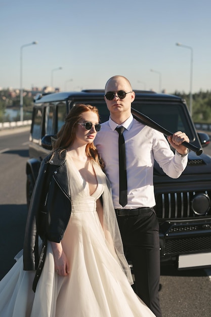 Casal brutal a noiva e o noivo com um morcego em jaquetas de couro perto do carro na estrada