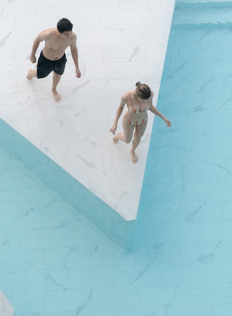 Casal caminhando para a piscina feita de ladrilho de pedra de mármore branco com água rasgada azul.