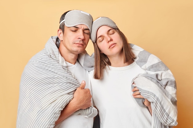 Casal cansado e exausto homem e mulher envoltos em cobertor isolados sobre um fundo bege precisam de descanso de pé com os olhos fechados sonolência