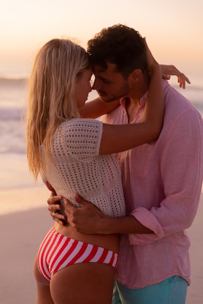 Foto casal caucasiano aproveitando o tempo na praia durante um lindo pôr do sol, abraçando e beijando com céu azul e mar ao fundo