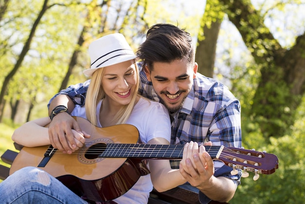 Casal caucasiano feliz sentado em um banco de parque, com ele tentando ensiná-la a tocar violão