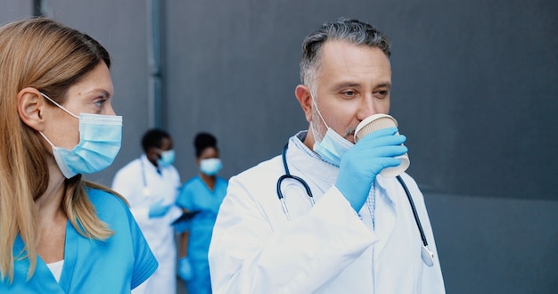 Foto casal caucasiano, homem e mulher, colegas médicos com máscaras médicas, caminhando, conversando e bebendo café para viagem. médicos masculinos e femininos discutindo trabalho e tomando uma bebida quente. médico e assistente.