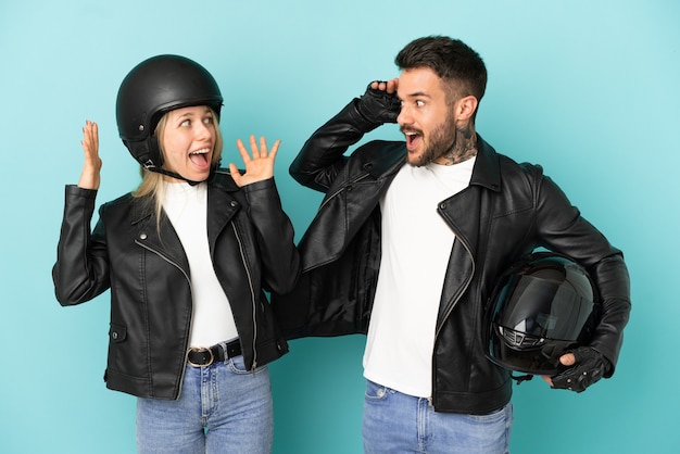 Casal com capacete de motociclista sobre fundo azul isolado com expressão facial de surpresa e choque
