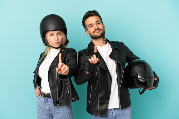 Casal com capacete de motociclista sobre fundo azul isolado, mostrando e levantando um dedo