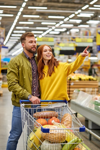 Foto casal com carrinho de compras comprando comida em mercearia ou supermercado, mulher aponta o dedo para o lado, pede ao marido para comprar algo