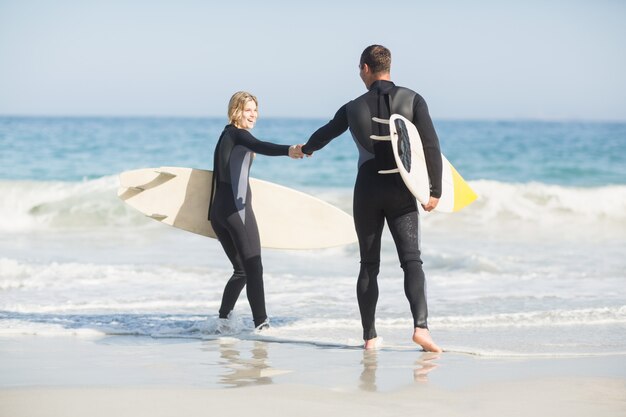 Casal com prancha de surf, segurando a mão na praia