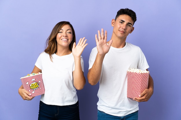 Casal comendo pipoca enquanto assiste a um filme roxo saudando com a mão e expressão feliz