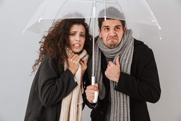 Casal congelado e chateado, vestindo roupas de outono, em pé sob um guarda-chuva isolado sobre uma parede cinza