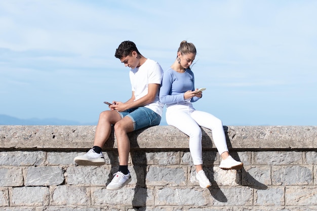 Casal de adolescentes tristes concentrados em seus telefones sentados em uma parede de pedra
