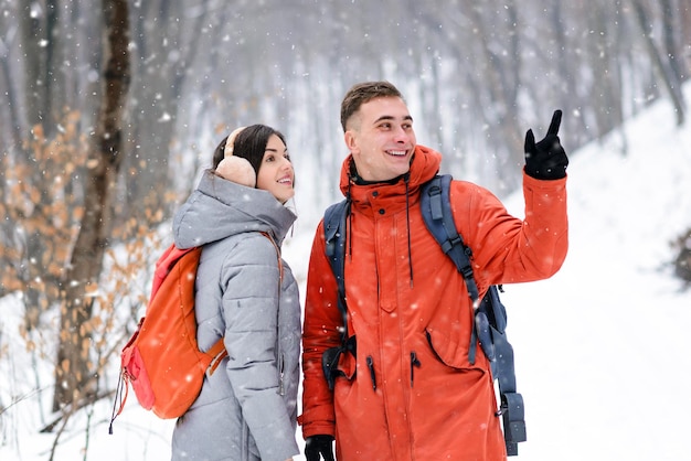 Casal de amigos com mochilas andando na floresta de neve de inverno, menino mostrando paisagens florestais de menina