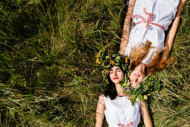 Casal de amigos de belas moças deitado na grama no verão e curtindo a vida. retrato de duas mulheres amorosas em vestidos tradicionais eslavos.