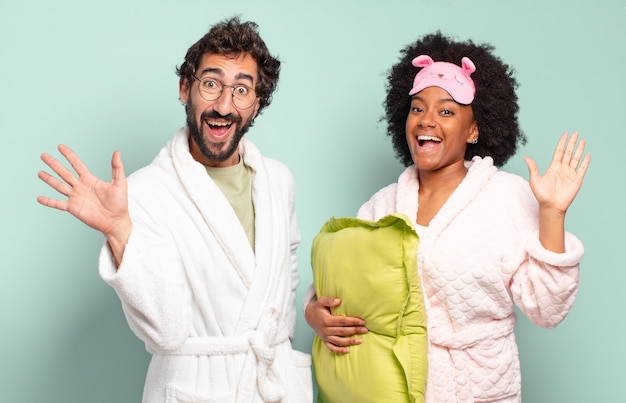 Casal de amigos multirraciais sorrindo alegremente e alegremente, acenando com a mão, dando as boas-vindas e cumprimentando você ou dizendo adeus. pijama e conceito de casa