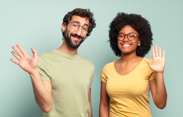 Casal de amigos multirraciais sorrindo feliz e alegre, acenando com a mão, dando as boas-vindas e cumprimentando você ou dizendo adeus