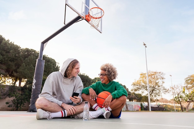 Casal de amigos rindo e se divertindo sentado na quadra depois de uma prática de basquete em um parque da cidade conceito de amizade e esporte urbano no espaço de cópia de rua para texto