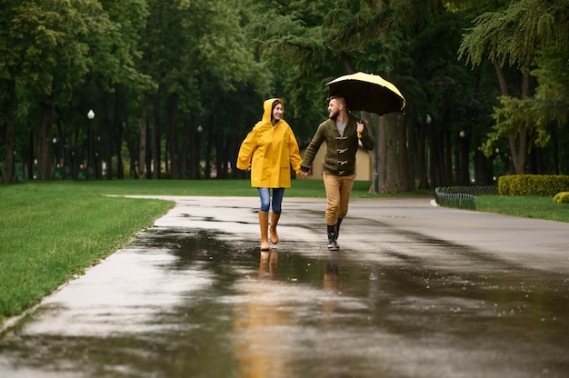 Casal de amor feliz corre no parque, dia chuvoso de verão. Homem e mulher sob o guarda-chuva na chuva, encontro romântico em uma trilha, tempo úmido no beco