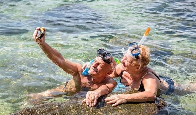 Casal de aposentados feliz tomando selfie em excursão no mar tropical com câmera de água e máscara de snorkel