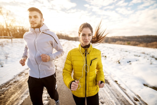 Casal de aptidão no sportswear de inverno correndo na natureza nevado com fones de ouvido.