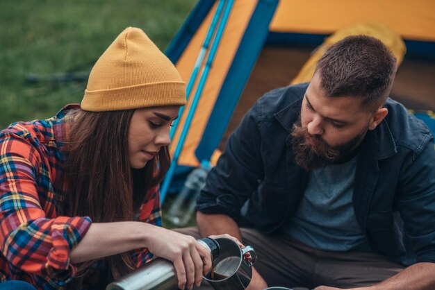 Foto casal de campistas bebendo café de uma xícara de metal e garrafa térmica