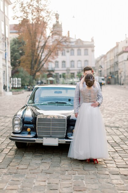 Casal de casamento de luxo feliz beijando e abraçando perto de carro retrô preto no antigo centro da cidade, edifícios antigos no fundo.