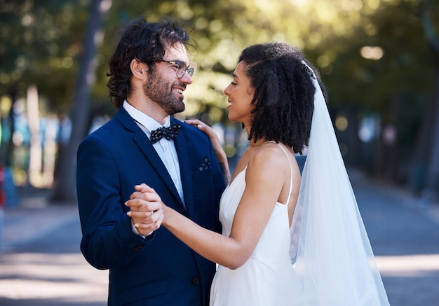 Casal de casamento inter-racial e dançando na rua com sorriso animado, felicidade ou futuro, mulher negra, noiva, homem e diversidade no casamento ao ar livre por amor, dança ou contato visual ao sol por árvores