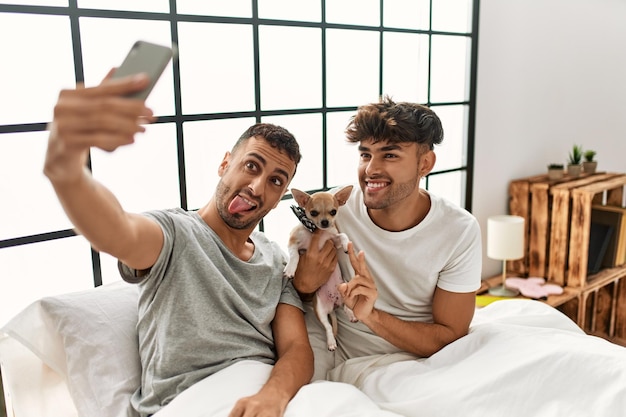 Casal de dois homens hispânicos faz selfie pelo smartphone sentado na cama com chihuahua no quarto