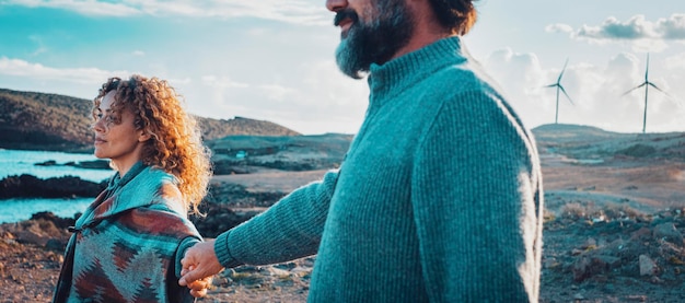 Casal de estilo de vida de viagem aprecia o pôr do sol juntos de mãos dadas e olhando para frente Amor e relacionamento no estilo de vida do viajante Wanderlust Coast oceano e moinhos de vento no fundo com céu azul Vida