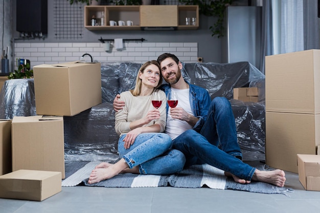 Casal de família apaixonado homem e mulher sentada no chão comemorando uma nova casa em um novo apartamento bebendo vinho entre caixas de papelão