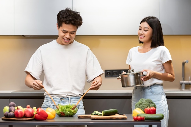 Casal de família asiática cozinhando, marido fazendo salada e esposa segurando a panela com refeição, desfrutando de preparação de jantar saudável juntos na cozinha moderna em casa. receitas de comida saborosa e conceito de nutrição