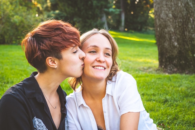 Casal de garotas apaixonadas se beijando, sentadas na grama em um parque em um dia ensolarado