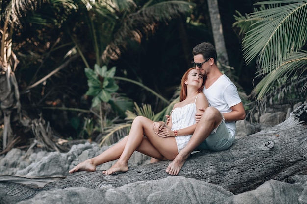 Casal de hipster na praia, garota ruiva em um vestido floral verde e homem com barba por fazer, sentado na velha grande árvore nua sem folhas. Phuket. Ã Â ¢ hailand