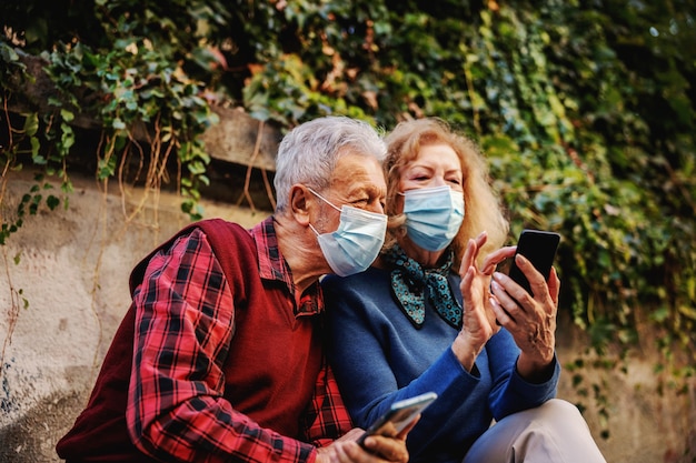 Casal de idosos apaixonado usando máscaras cirúrgicas protetoras e olhando para um telefone inteligente