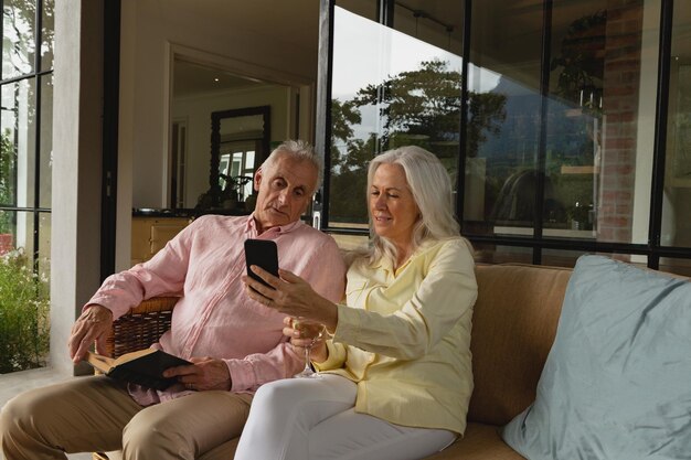 Foto casal de idosos ativos usando telefone celular e lendo um livro na varanda