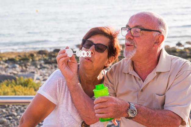 Casal de idosos casados sentados na praia soprando uma bolha de sabão com o mar ao fundo - se divertindo juntos