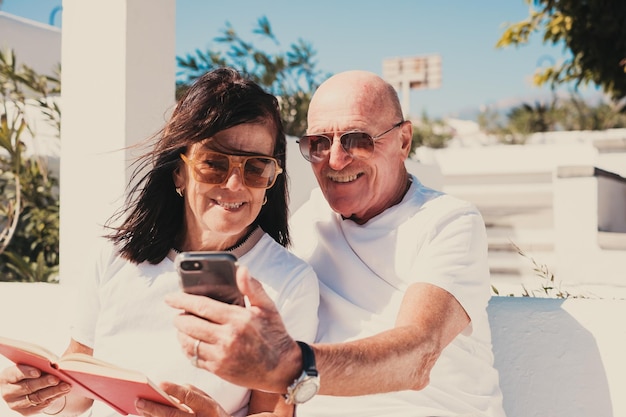 Casal de idosos caucasianos felizes relaxando sentados ao ar livre em um banco com um livro e um telefone celular