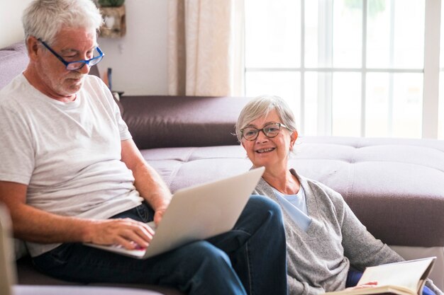 Casal de idosos em casa - homem com laptop e óculos sentado no sofá - mulher sentada no chão lendo um livro - conceito interior e amor