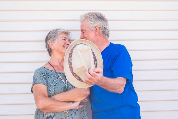 Casal de idosos feliz sorrindo com chapéu na mão em um dia ensolarado com parede de madeira branca - idosos alegres aproveitam o estilo de vida com amor e diversão juntos