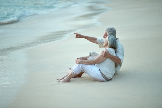 Casal de idosos felizes descansando em uma praia tropical, homem apontando pela mão