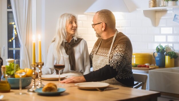 Casal de idosos felizes e apaixonados têm uma noite romântica cozinhando e jantando na cozinha celebrando aniversário idosos têm uma tarde romântica com vinho mesa festiva no interior aconchegante da cozinha