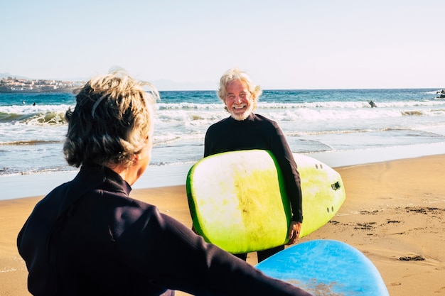 Casal de idosos na praia com roupas de neoprene segurando uma mesa de surf pronta para surfar na praia - adultos ativos e aposentados fazendo atividades felizes juntos em suas férias ou tempo livre