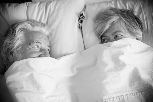 Casal de idosos olhando e sorrindo com amor e afeto debaixo do cobertor na cama - adultos aposentados e maduros casados no quarto pela manhã antes de se levantar