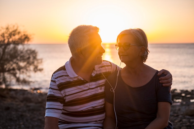 Foto casal de idosos passeando na praia com o pôr do sol - casal aposentado ouvindo música com os mesmos fones de ouvido