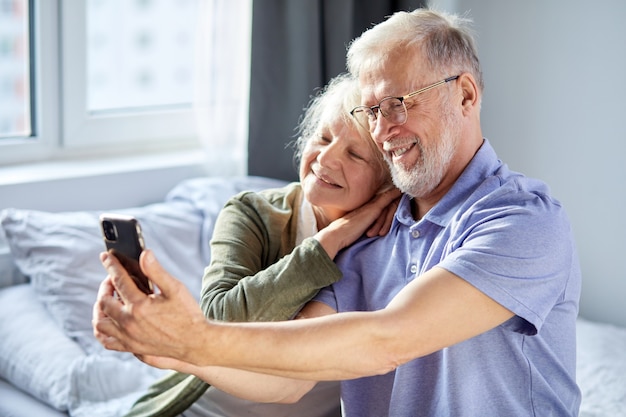 Casal de idosos tirando foto no smartphone, enquanto está sentado no quarto, sente-se sorrindo. conceito de tecnologia de estilo de vida de sociedade de pessoas sênior. homem e mulher compartilham mídias sociais juntos no bem-estar doméstico