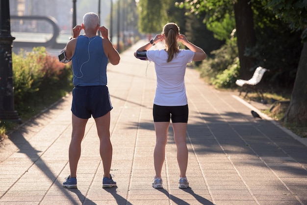 casal de jogging verifica a lista de reprodução de música no telefone e planeja a rota antes do treino matinal com o nascer do sol na cidade e o reflexo do sol ao fundo