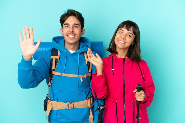 Casal de jovens alpinistas isolados em fundo azul saudando com a mão com expressão feliz