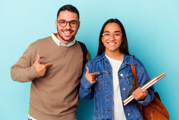 Casal de jovens estudantes de raça mista isolado em fundo azul pessoa apontando com a mão para um espaço de cópia de camisa, orgulhoso e confiante