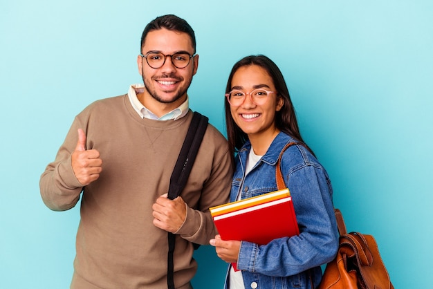 Casal de jovens estudantes de raça mista isolado em fundo azul, sorrindo e levantando o polegar