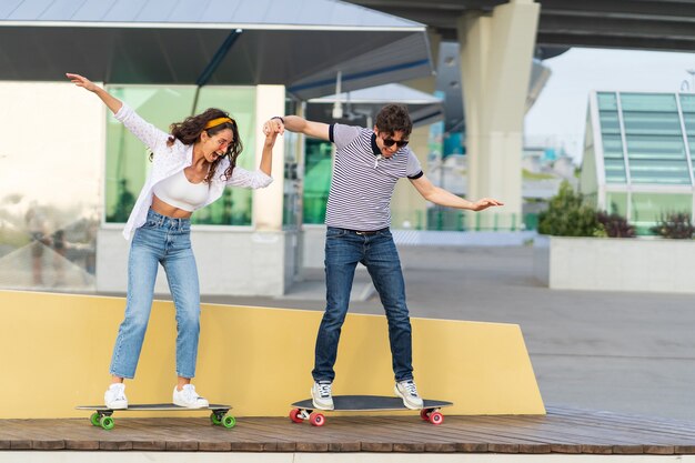 Casal de jovens patinadores ativos aprendem a andar de longboard juntos de mãos dadas e rindo em pé de skate