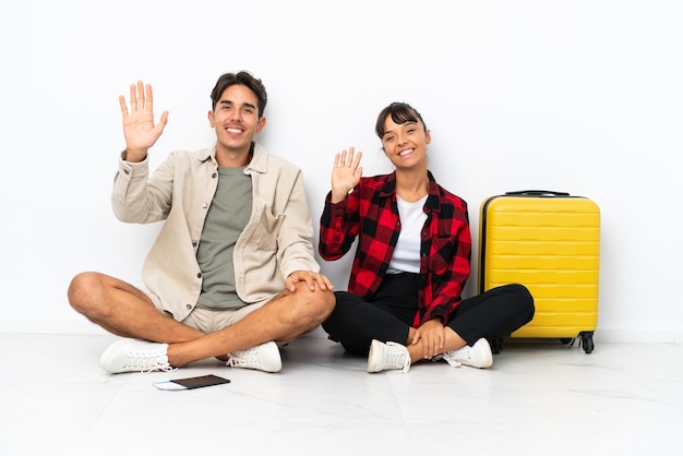 Casal de jovens viajantes de raça mista sentados no chão isolados no fundo branco saudando com a mão com expressão feliz