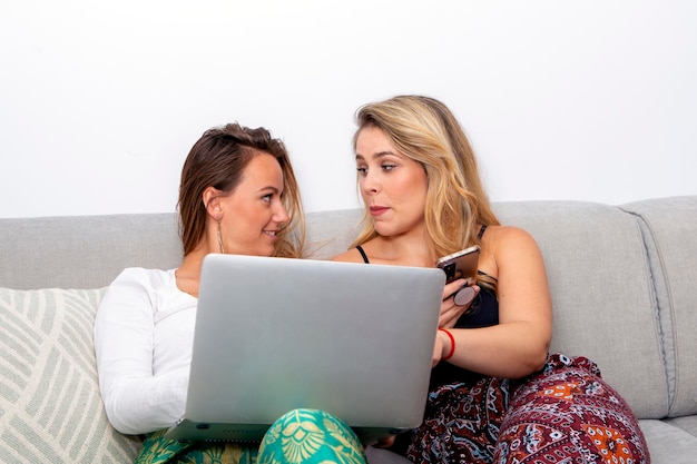 Casal de lésbicas sorridente no sofá olhando para laptop e telefone inteligente